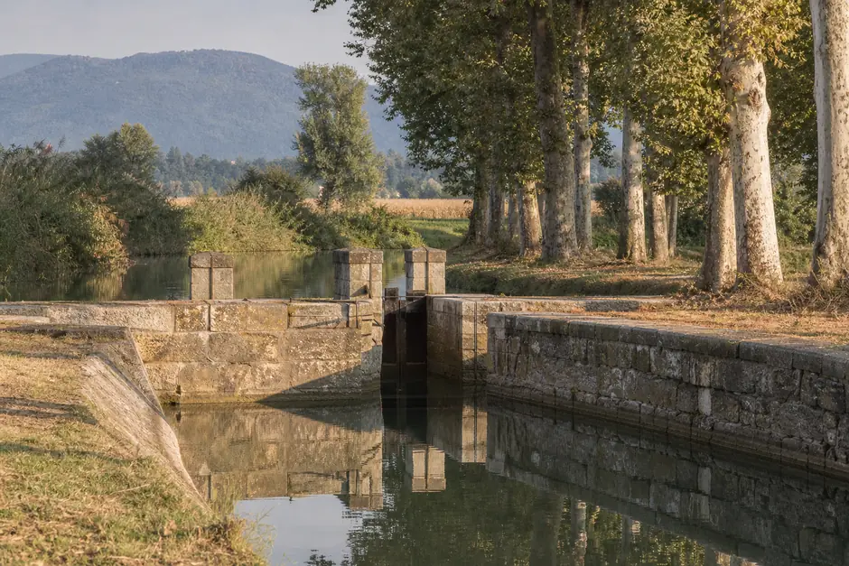 Canale d'irrigazione con chiusa in pietra in una campagna pianeggiante, pioppi lungo la sponda, acqua ferma (immagine generata con AI)
