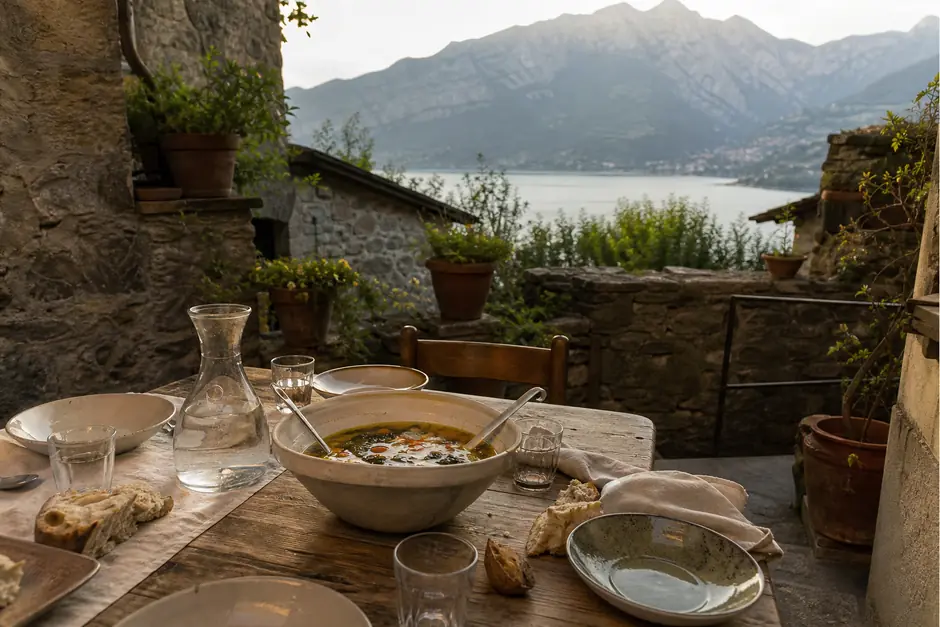 Tavola rustica apparecchiata in un cortile di pietra con una zuppiera di verdure e pane spezzato (immagine generata con AI)