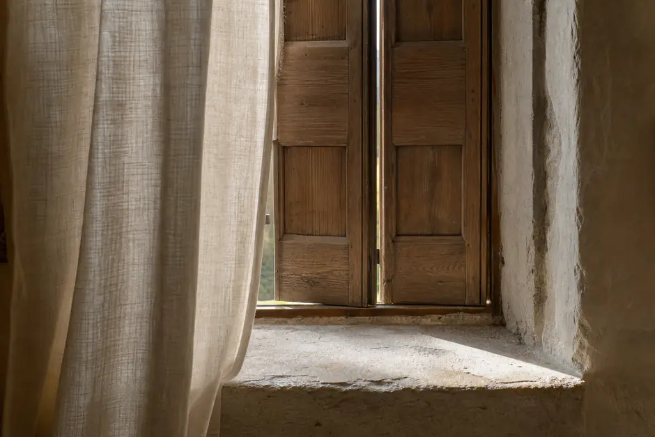 Finestra di una casa in pietra con scuri di legno socchiusi e una tenda di lino tirata a meta (immagine generata con AI)