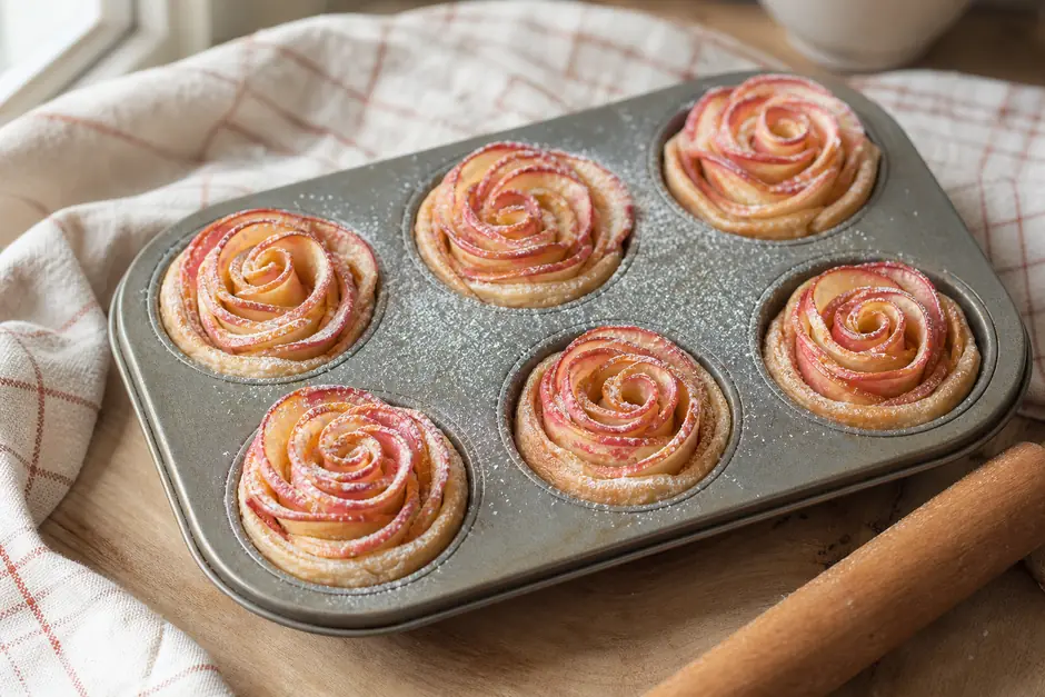 Sei rose di mele in una teglia da muffin, spolverate di zucchero a velo (immagine generata con AI)