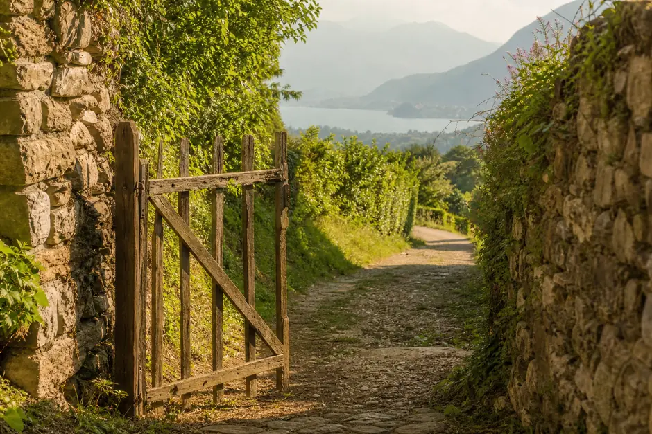 Vecchio cancello di legno aperto su una strada di campagna, colline e lago sullo sfondo (immagine generata con AI)